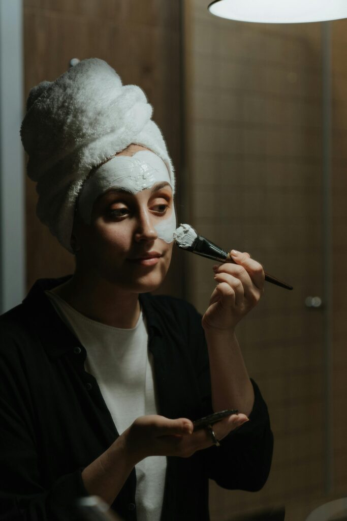 A woman in a towel applies a face mask as part of her skincare routine in a cozy setting.